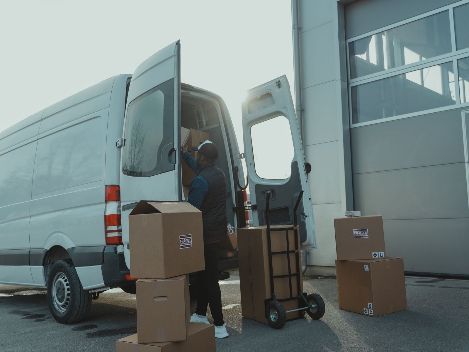 A worker loading sealed cartons into the back of an unmarked white cargo truck at a warehouse bay, hand truck and stacked boxes waiting on the pavement